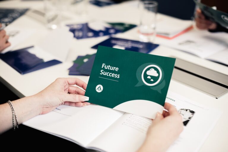 A close up of a person's hands, holding a branded print out that reads 'future success'. On the table in the background is a notebook and various other print outs to represent a workshop setting.