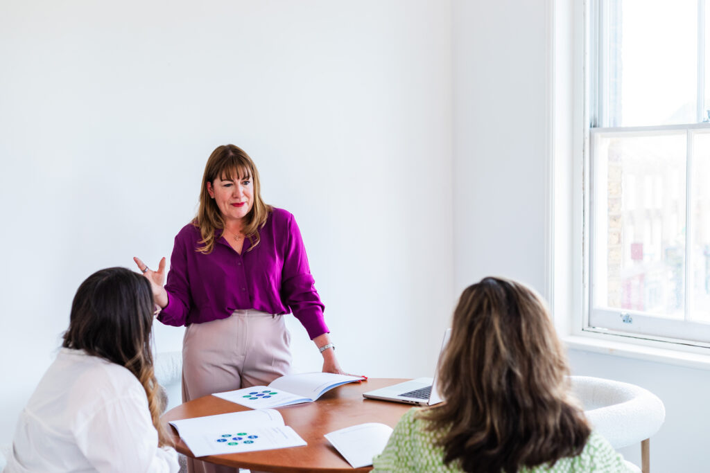 Antoinette Oglethorpe in a purple blouse stands and gestures while speaking to two seated colleagues at a round table with documents and a laptop in a bright, white office room.