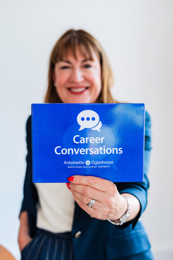 Antoinette Oglethorpe smiling holding a purple Career Conversations Toolkit that reads "Career Conversations Antoinette Oglethorpe" with a speech bubble graphic, against a plain light background.
