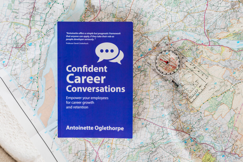 A purple book titled "Confident Career Conversations" by Antoinette Oglethorpe sits on an unfolded map next to a clear compass. The cover promotes employee growth and retention.