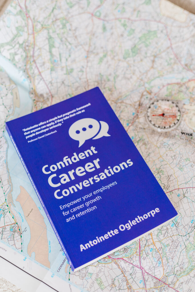 A purple book titled "Confident Career Conversations" by Antoinette Oglethorpe rests on a map next to a compass. The book's subtitle reads, "Empower your employees for career growth and retention.