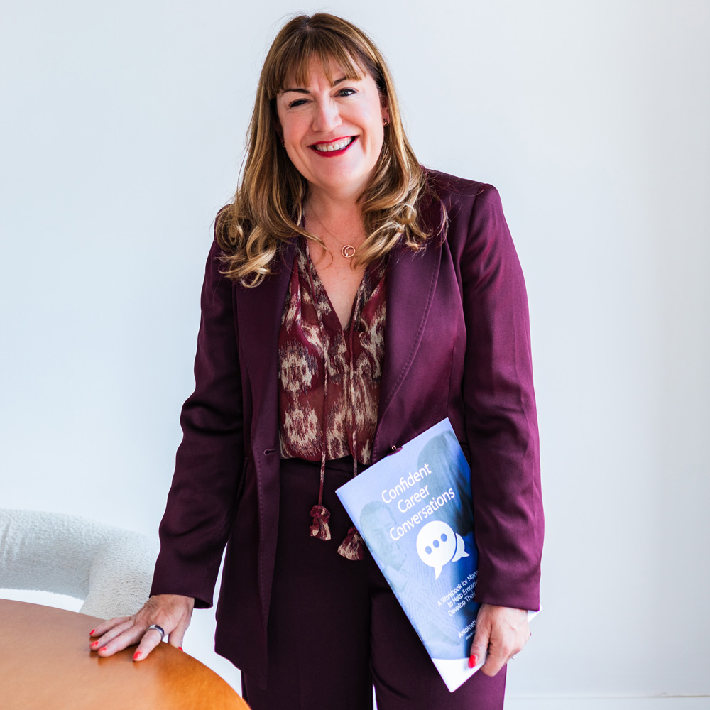 Antoinette Oglethorpe keynote speaker and author in a burgundy suit stands by a table, holding a booklet titled "Confident Career Conversations" against a plain white background.