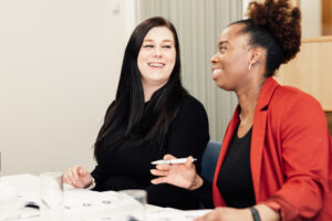 Two women sit at a table, smiling and talking. One woman has long dark hair and wears a black top, whilst the other has curly hair pulled back and wears a red blazer. Papers and glasses are on the table in front of them.