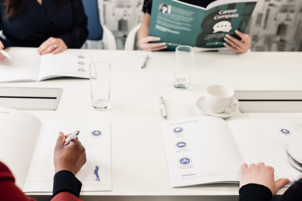Three people sit at a table with open notebooks, pens, and glasses of water. One person reads a green workbook titled “Career Compass.” The scene suggests a professional meeting or workshop.