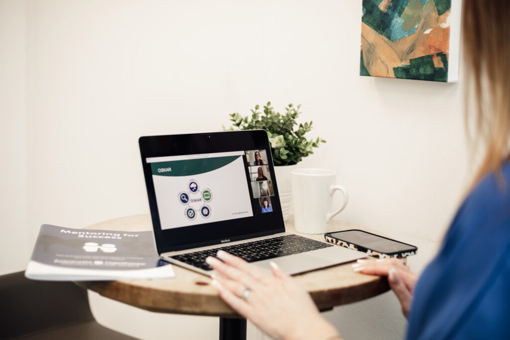 A person sits at a small round table, participating in a video call on a laptop. The screen displays several people and a chart. A phone, coffee mug, booklet, and plant are on the table. A painting hangs on the wall.