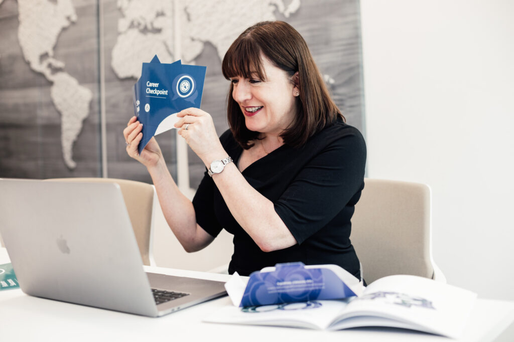 Antoinette Oglethorpe sits at a desk smiling whilst holding up a card from her Career Conversation Toolkit titled "Career Checkpoint" in front of her laptop delivering a masterclass. Open books and papers are spread out on the table. A world map is visible in the background.