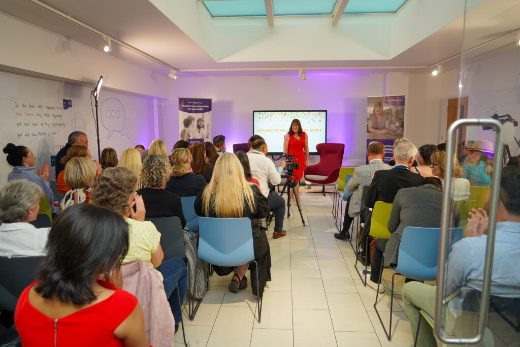 Antoinette Oglethorpe stands and speaks in front of an audience seated in a modern, brightly lit room with colourful chairs, a large screen, and posters on the walls at her book launch party celebrating her Amazon #1 best seller Confident Career Conversations.