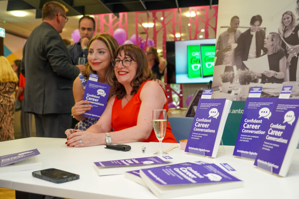 Antoinette Oglethorpe and another woman sit at a table smiling for a photo at a book signing event. Books titled “Confident Career Conversations” are displayed on the table along with a glass of champagne and a mobile phone. Other people are in the background.