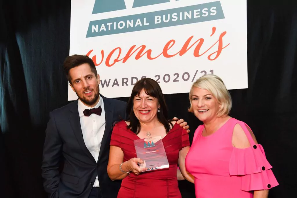 Three people pose and smile at the National Business Women's Awards 2020/21; the woman in the centre holds a glass award, with two others standing on either side under an event sign.