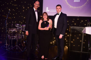 Three people in formal attire stand on stage at an awards event. Antoinette Oglethorpe in the centre is holding a trophy and smiling. Behind them are drums, a lectern, and a backdrop with twinkling lights and event branding.