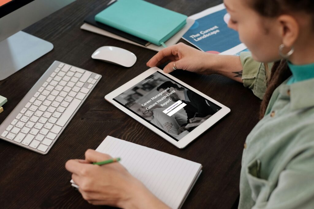 A person sitting at a desk uses a tablet displaying a personalised report from the career management culture health check, with a keyboard, mouse, notebook, pencil, and documents nearby.