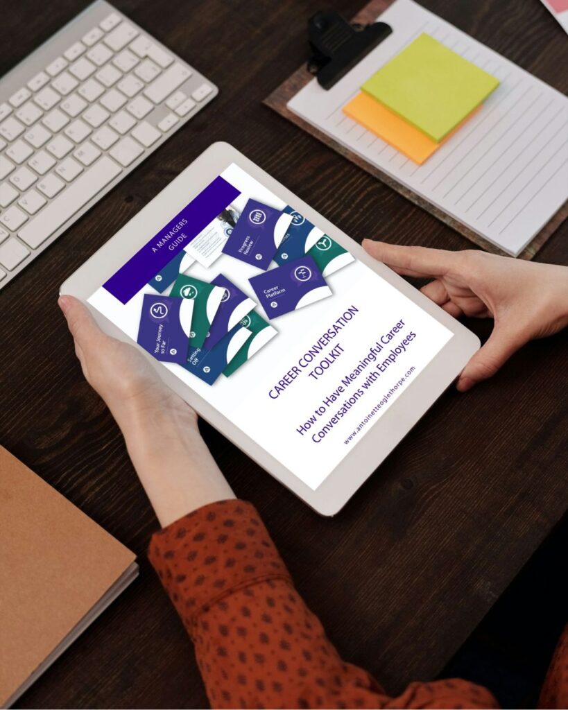 A person holding a tablet displaying a document titled " A Managers Guided to the Career Conversation Toolkit" at a desk with a keyboard, notepad, clipboard, and closed notebook.