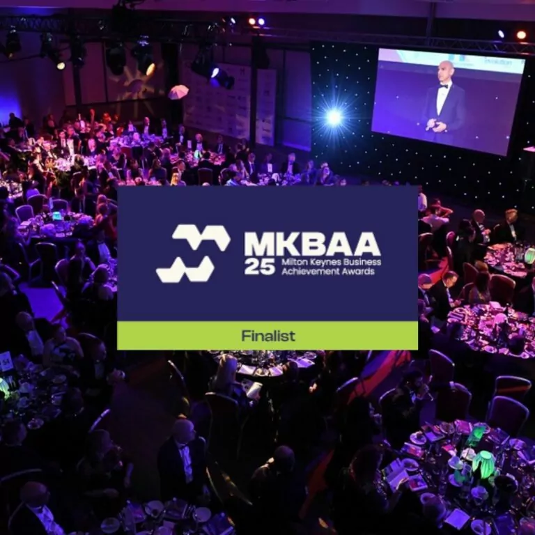 A large formal event with round tables full of guests in evening dress, a stage with a speaker, and a big screen. Overlaid text reads "MKBAA 25 Milton Keynes Business Achievement Awards - Finalist.