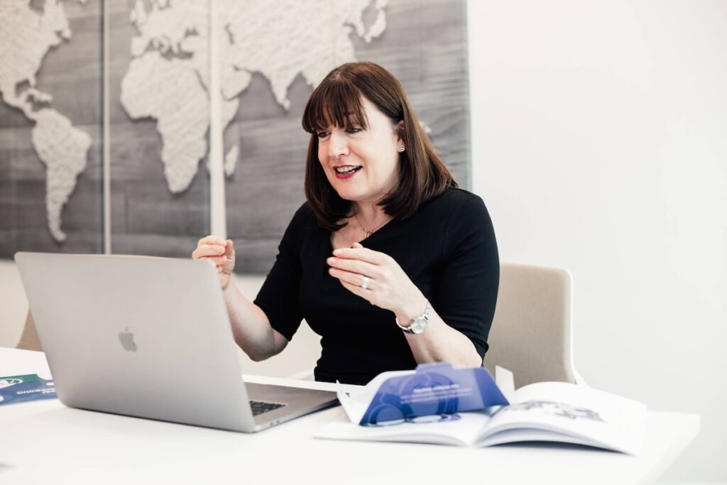 Antoinette Oglethorpe with shoulder-length brown hair sits at a desk, gesturing as she talks to her laptop delivering a virtual session as part of the Certified Facilitator Programme. An open book and papers are on the desk, and a world map is displayed on the wall behind her.