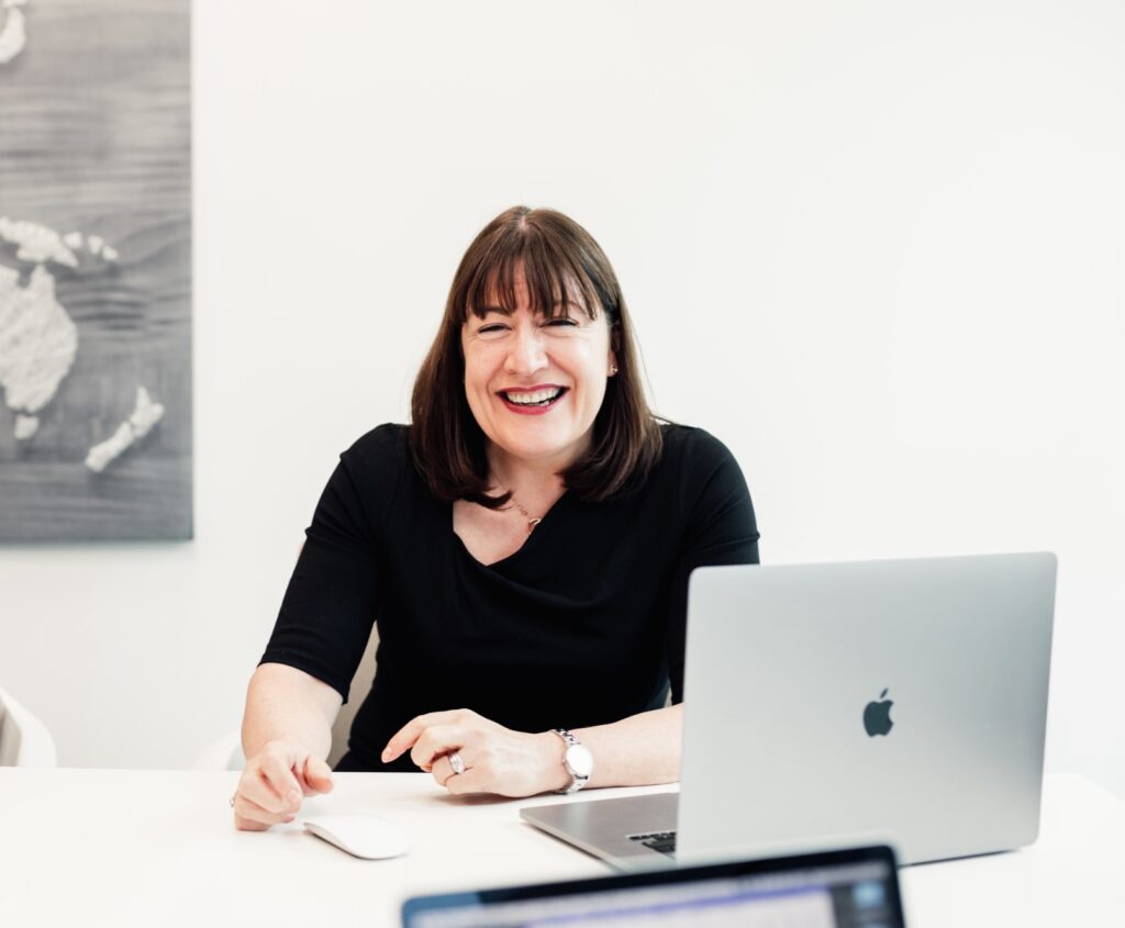 Antoinette Oglethorpe is in a black top and is smiling whilst sitting at a desk with an open laptop. A map is on the wall behind her, she has just delivered the webinar how to design and implement a mentoring programme that maintains momentum