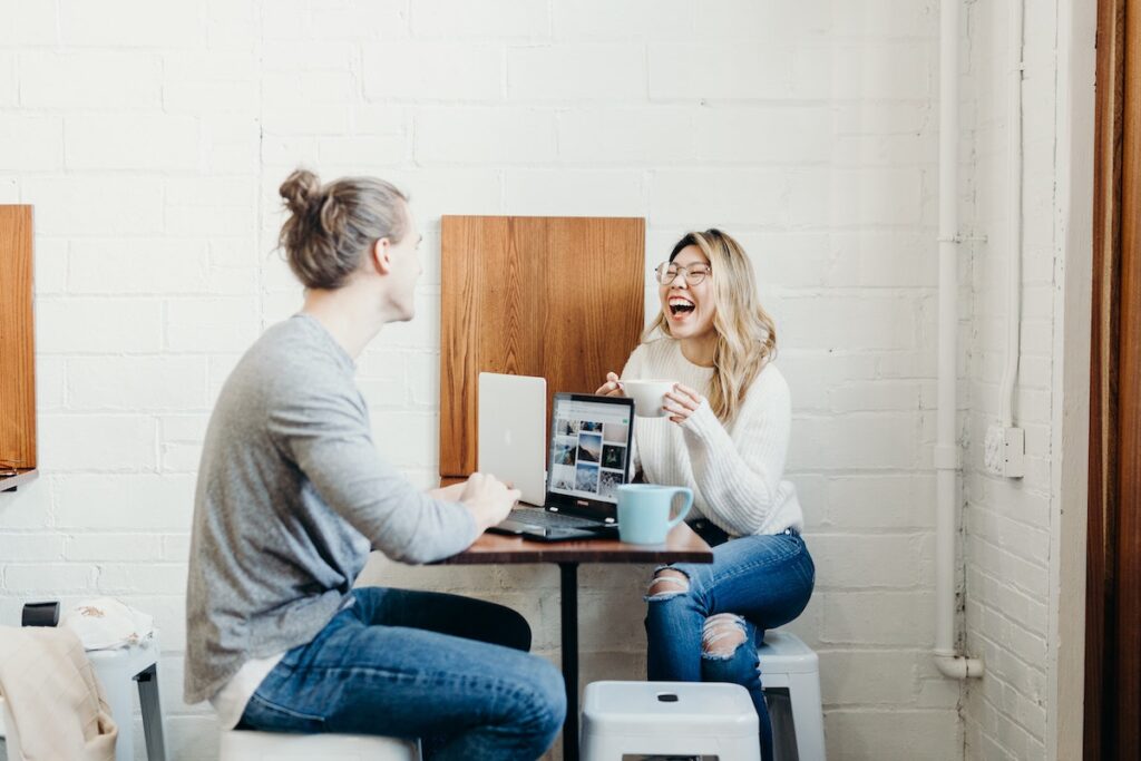 A man and a woman sit opposite each other at a small table with a laptop and mugs, laughing and having a great career conversation in a lively, casual modern café setting.