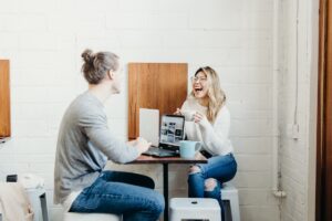 A man and a woman sit opposite each other at a small table with a laptop and mugs, laughing and having a great career conversation in a lively, casual modern café setting.