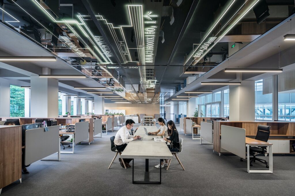 Three people sit at a table in a modern, open-plan office with large windows, wooden cubicles, and ceiling lights, discussing career management and mentoring challenges. The space is bright and organised with mirrored panels featuring white graphic designs.