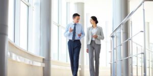A man and a woman in business attire walk and talk in a bright, modern office corridor with large windows and metal railings, engaging in effective career conversations.
