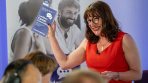 Antoinette Oglethorpe in a red dress holds up her best selling book titled "Confident Career Conversations" whilst speaking to an audience. A banner with people and speech bubbles is visible in the background.