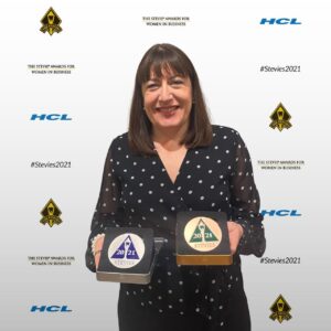 A smiling woman in a black polka-dot blouse proudly holds two awards, posing in front of the 2021 Stevie Awards for Women in Business step-and-repeat backdrop.