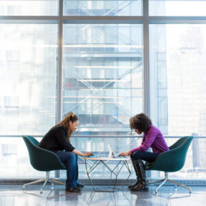 Two people sit facing each other at a small round table with laptops open, collaborating on their career goals in a modern office space with large glass windows revealing an urban cityscape in the background.