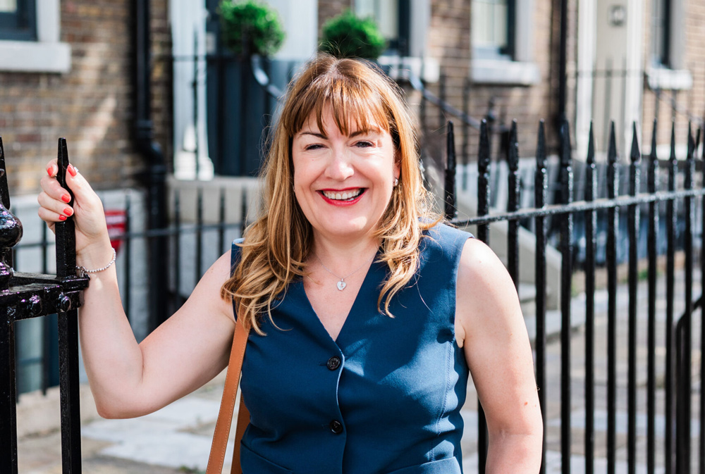 Antoinette Oglethorpe with light brown hair and a fringe, wearing a sleeveless blue dress, stands smiling by a black iron fence on a sunny city street. She holds the fence with one hand and has a tan shoulder bag.
