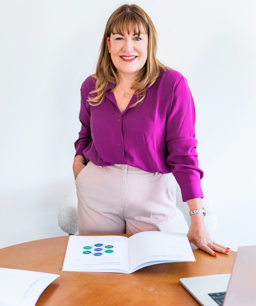 Antoinette Oglethorpe wearing a purple blouse and light trousers stands by a round table, smiling. An open career compass workbook with charts and a laptop are on the table in front of her. The background is bright and minimal. She is prepapring to delivery her career compass masterclass.