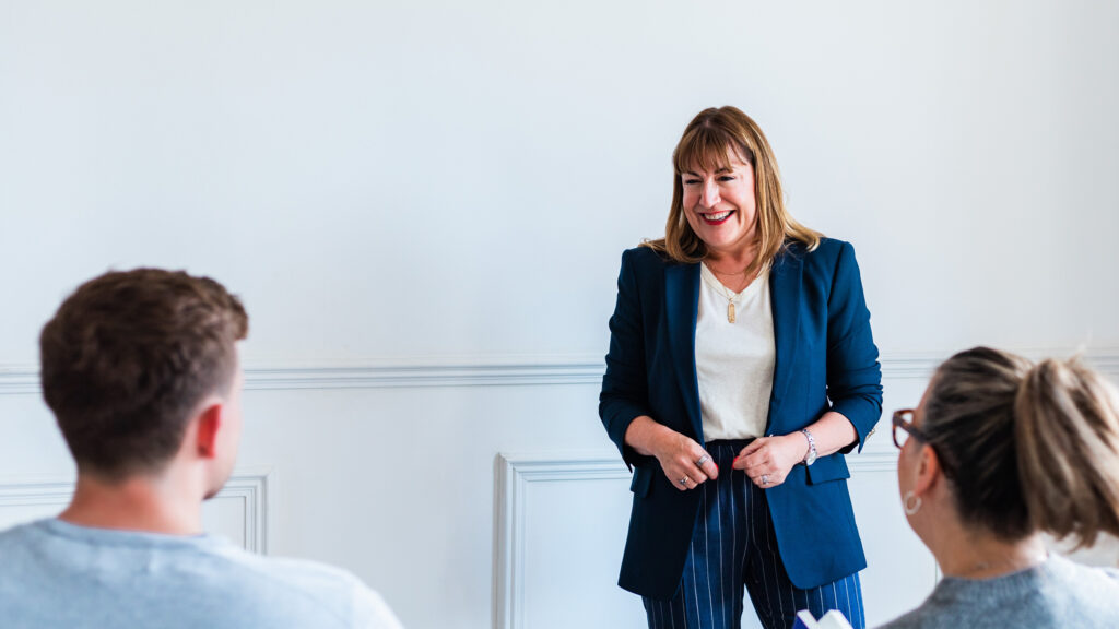 Antoinette Oglethorpe keynote speaker and global authority on career development, leadership and the future of work in a blue blazer stands and smiles whilst speaking to seated people in a bright room with a white wall.