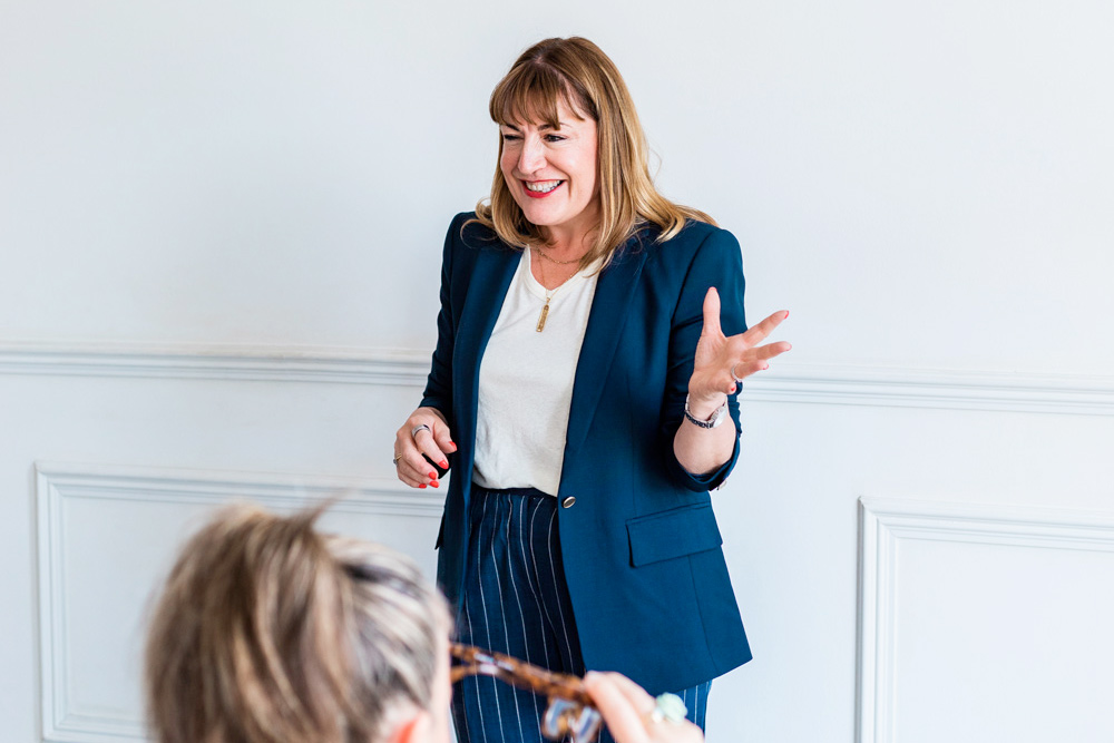 Antoinette Oglethorpe in a navy blazer and striped trousers is smiling and gesturing whilst delivering a bespoke masterclass to people in a bright, white room.