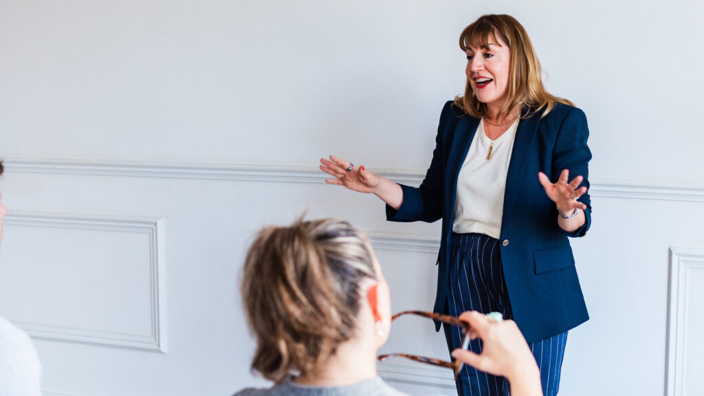 Antoinette Oglethorpe career management & leadership speaker stood in a navy blazer and striped trousers stands and gestures while delivering a keynote with two seated people in a bright room with white walls, engaged in what she's saying.