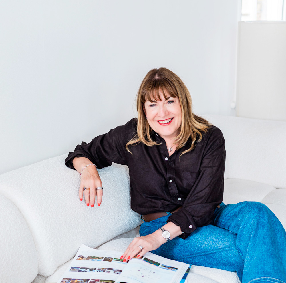 Antoinette Oglethorpe with straight light brown hair and a fringe, wearing a black shirt and blue jeans, sits smiling on a white sofa with a magazine open in front of her.