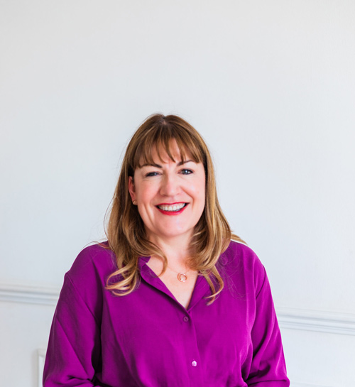 Antoinette Oglethorpe headshot, she has straight, light brown hair and a fringe is smiling at the camera. She is wearing a bright purple blouse and a necklace, standing against a plain white background.