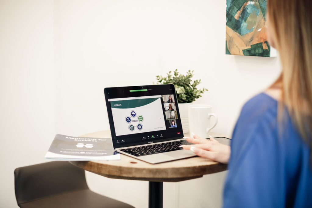 A woman sits at a small round table, attending a webinar on-demand on her laptop. A booklet, white mug, and small plant are on the table, and a painting hangs on the white wall in the background.