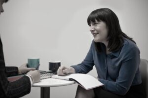 Two people sit at a small round table with notebooks, pens, and mugs. A woman in a blue blouse smiles whilst talking to the other person, who is writing in a notebook.