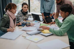 Four people sit and one stands around a table covered in notebooks, folders, and sticky notes, collaborating and discussing work. Laptops and pens are visible, showing their dedication to group study or team projects focused on operational excellence.