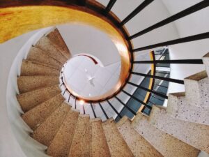 A spiral staircase with beige steps and black railings, viewed from above. The curved handrail is light brown, and the stairwell forms a circular pattern—resembling career progression—leading down to a white floor and ceiling fan.