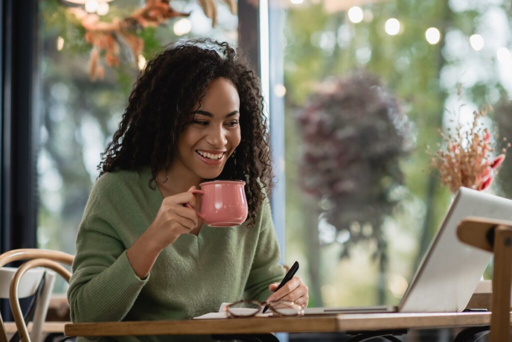 A woman with curly hair, wearing a green jumper, smiles whilst holding a pink mug and looking at a laptop. She sits at a wooden table with a notebook and pen, ready to learn the skills needed for the future workplace.