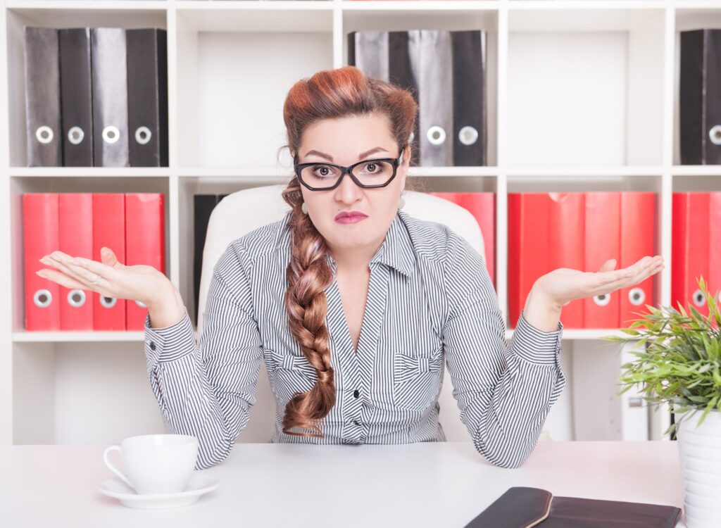 A woman with glasses and a long plait sits at a desk, shrugging with both hands raised. She looks confused or uncertain. Behind her are shelves with red and black files. A plant and a cup are on the desk.