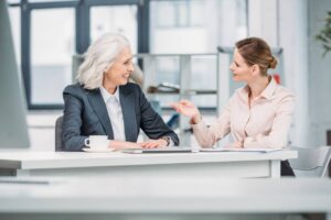Two women in business attire sit at a desk, engaged in friendly career development conversations. One has grey hair and a suit, the other has brown hair in a bun and a light blouse. A coffee cup and laptop are on the desk.