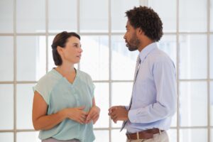 Two people stand indoors, facing each other and having a conversation. One is a woman in a light blue blouse, the other a man in a light blue shirt and tie. They appear engaged in discussing career development questions to ask employees.