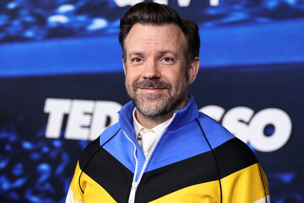 A man with short brown hair and a beard smiles at the camera, wearing a colourful jacket with blue, black, yellow, and white stripes. The blurred background features large white text reading 