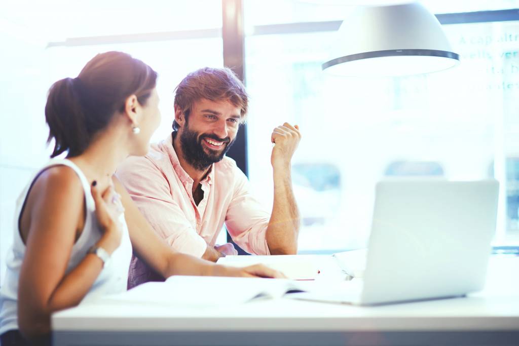 Two people sit at a table in a bright office, smiling and talking whilst looking at a laptop. Papers and a notebook are spread out in front of them as they share career conversation tips beneath a large hanging lamp.