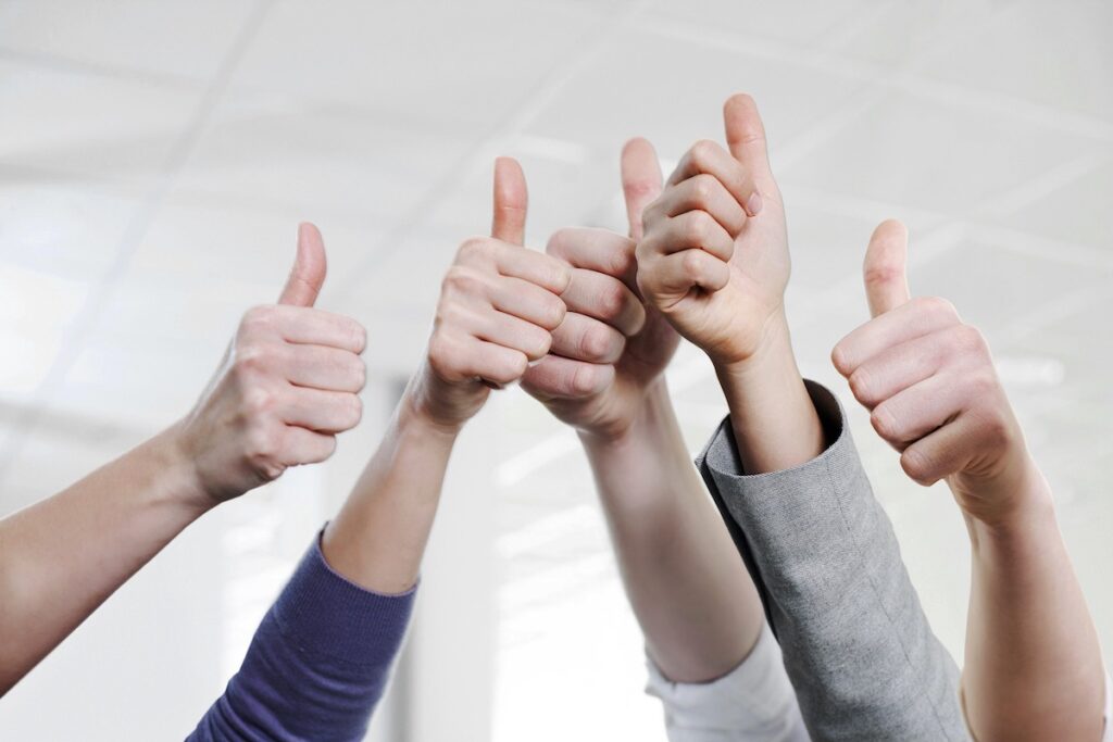 Five hands giving a thumbs-up gesture, raised against a bright, blurred indoor background, suggesting approval or agreement.