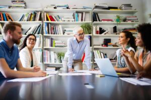 Five people sit around a table in a bright office, discussing and smiling, with open laptops and papers in front of them as they work together to develop staff. Bookshelves filled with books and plants are in the background.