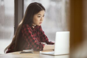 A young woman with long dark hair in a red checked shirt sits at a table by the window, focused on her laptop and notebook as she prepares for a Mentoring Programme Engagement.
