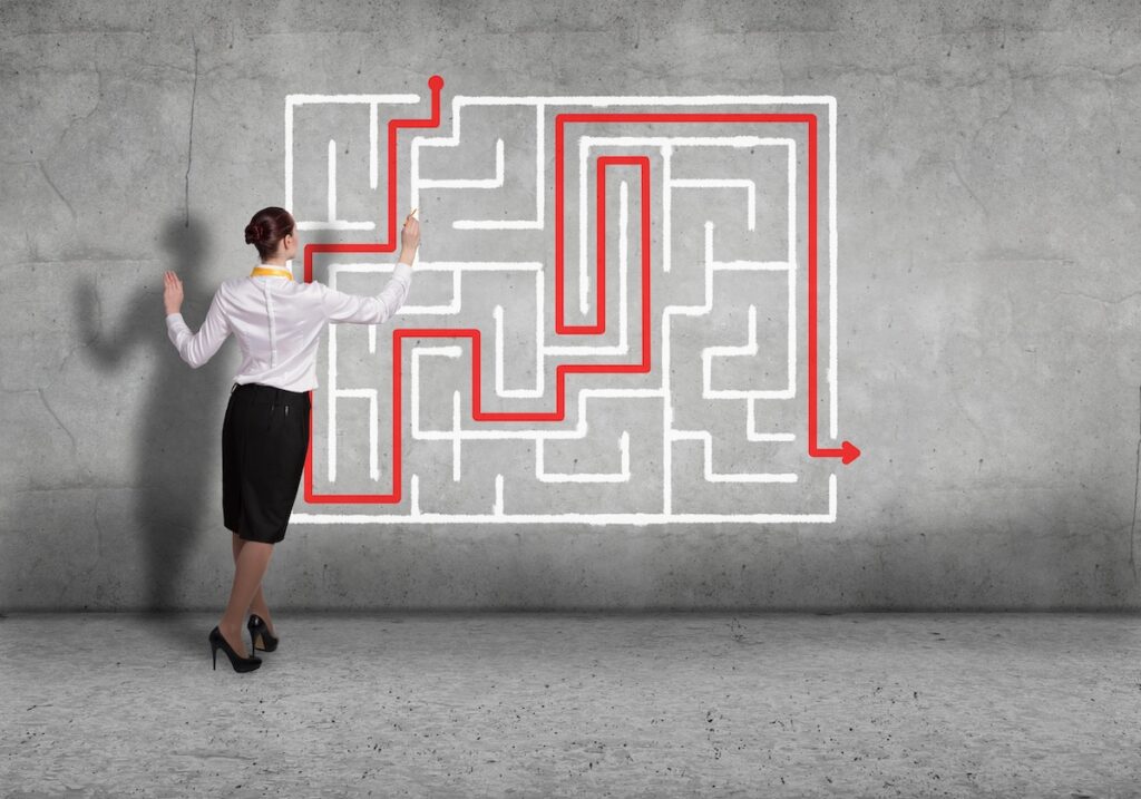 A woman in business attire stands before a wall with a drawn maze, tracing a red solution path—symbolising the strategic navigation of High-Potential Talent from entrance to exit with her finger.