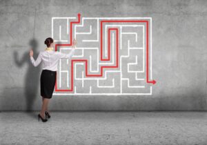 A woman in business attire stands before a wall with a drawn maze, tracing a red solution path—symbolising the strategic navigation of High-Potential Talent from entrance to exit with her finger.