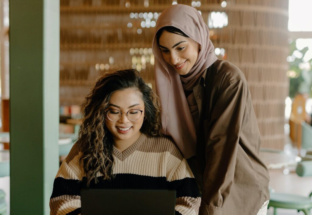 Two women smile while looking at a laptop together in a cosy indoor setting, showing great EQ. One woman wears glasses and a striped jumper, while the other, in a light pink hijab and brown shirt, stands beside her.