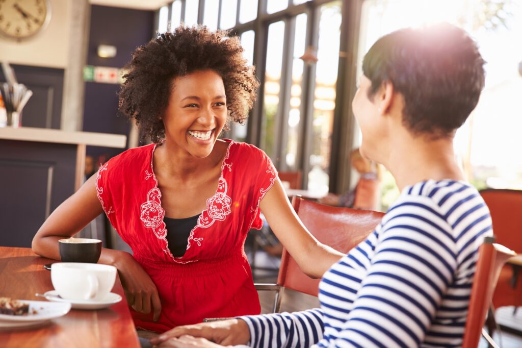 Two women, both managers, sit at a table in a bright café, smiling and talking. Sunlight streams through large windows as they discuss strategies to boost growth, creating a warm, cheerful atmosphere.
